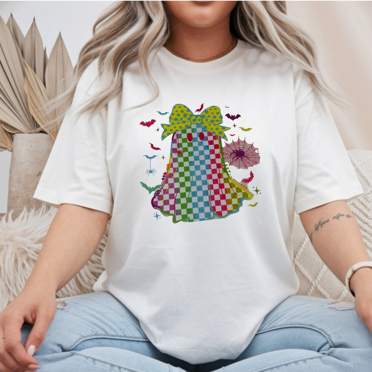 White Gildan Softstyle tee mockup with a rainbow checkered ghost in a polka dot bow, surrounded by bats and a spiderweb