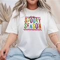 White Gildan Softstyle tee mockup with distressed rainbow SPOOKY SEASON letters, a purple bow, and red checkerboard borders