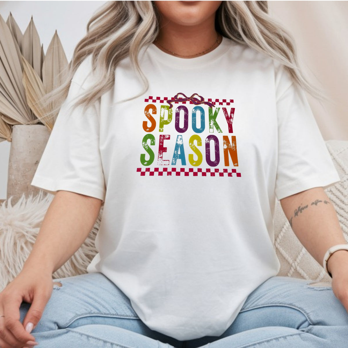 White Gildan Softstyle tee mockup with distressed rainbow SPOOKY SEASON letters, a purple bow, and red checkerboard borders